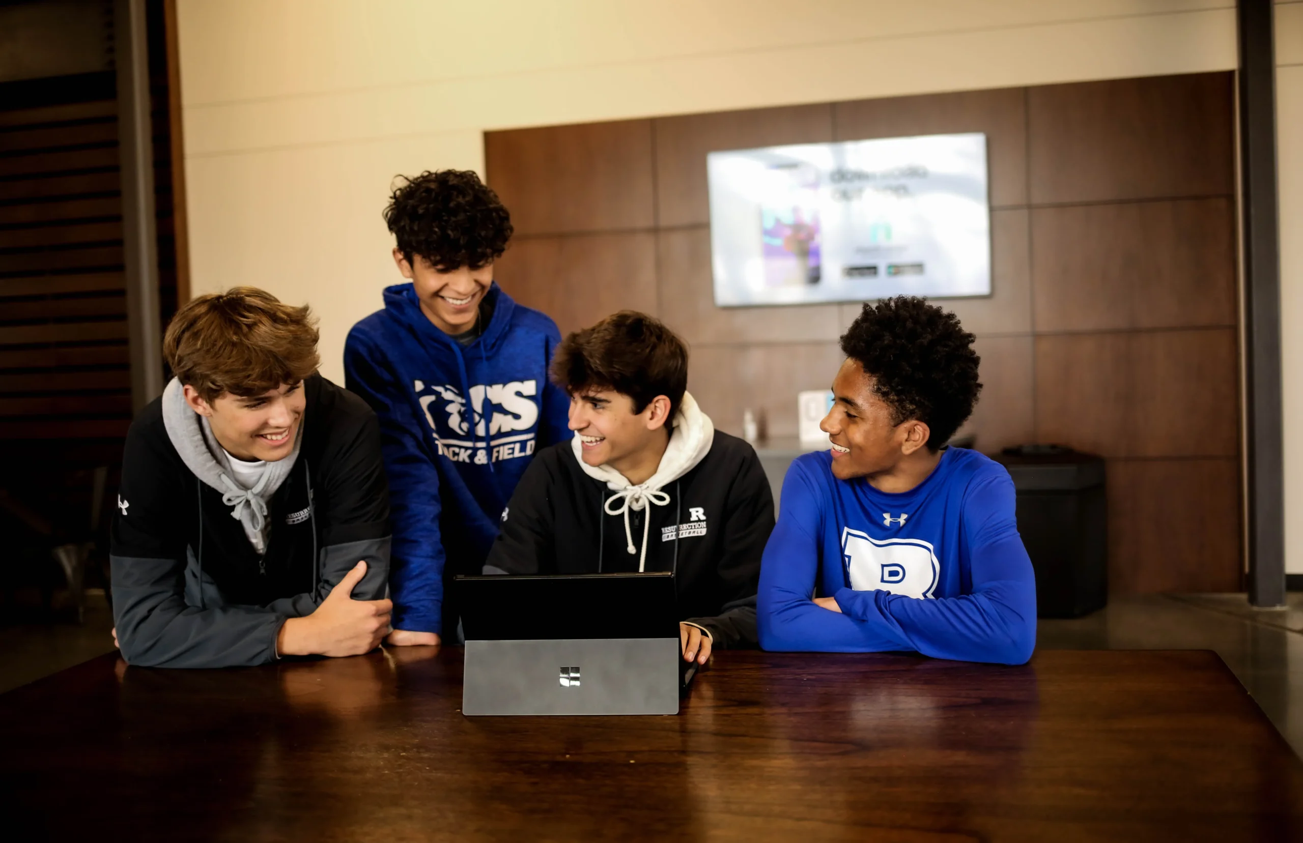 Four young men sit together around a laptop at a wooden table, smiling during a casual group session.