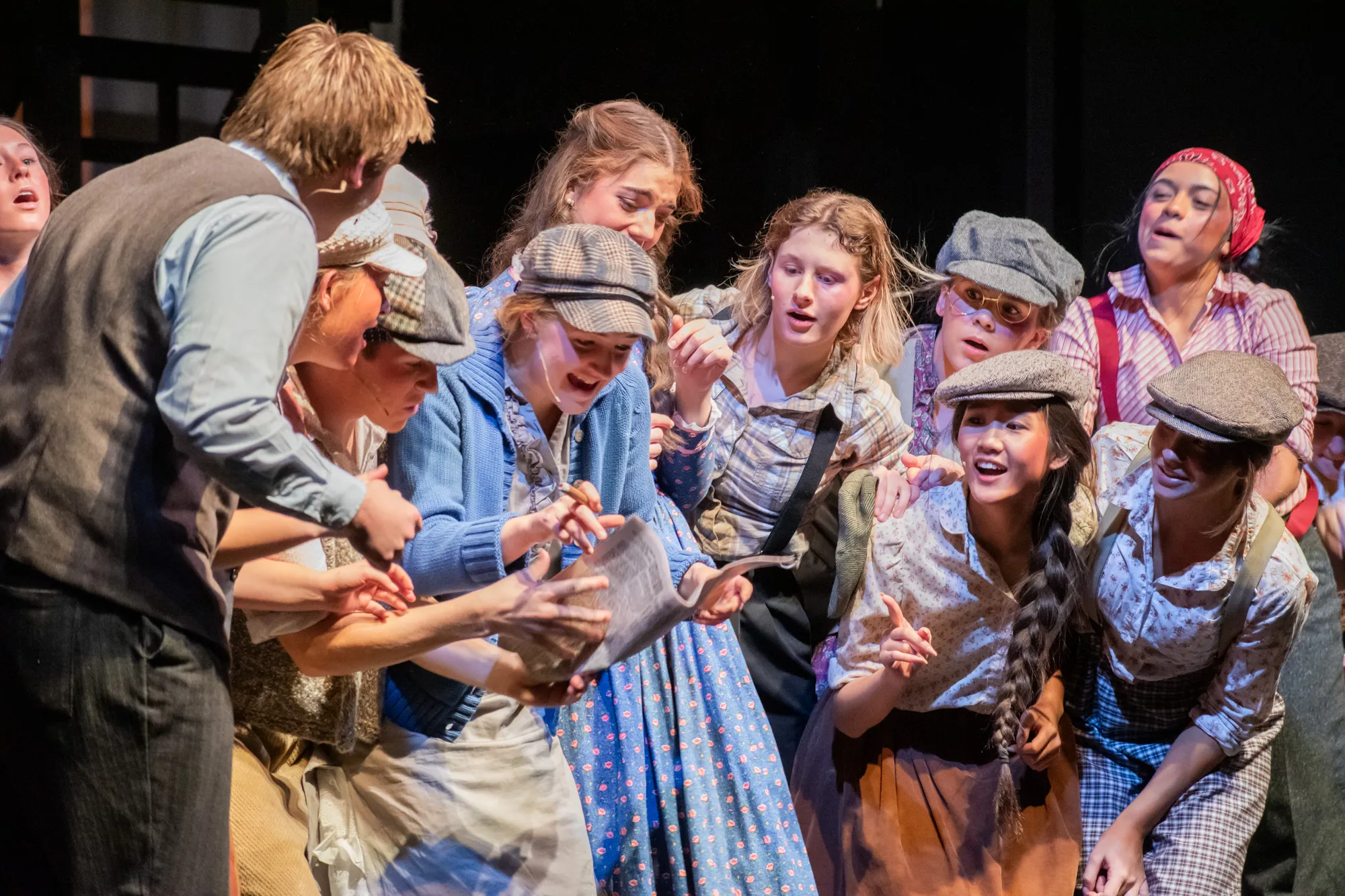 A crowded group of young performers in period costumes lean together onstage, reacting excitedly as one holds up a newspaper.