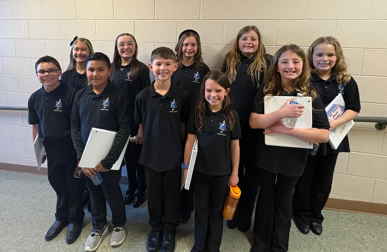 Ten students in matching black Bella Voce polos stand together in a school hallway, with several holding binders and water bottles.