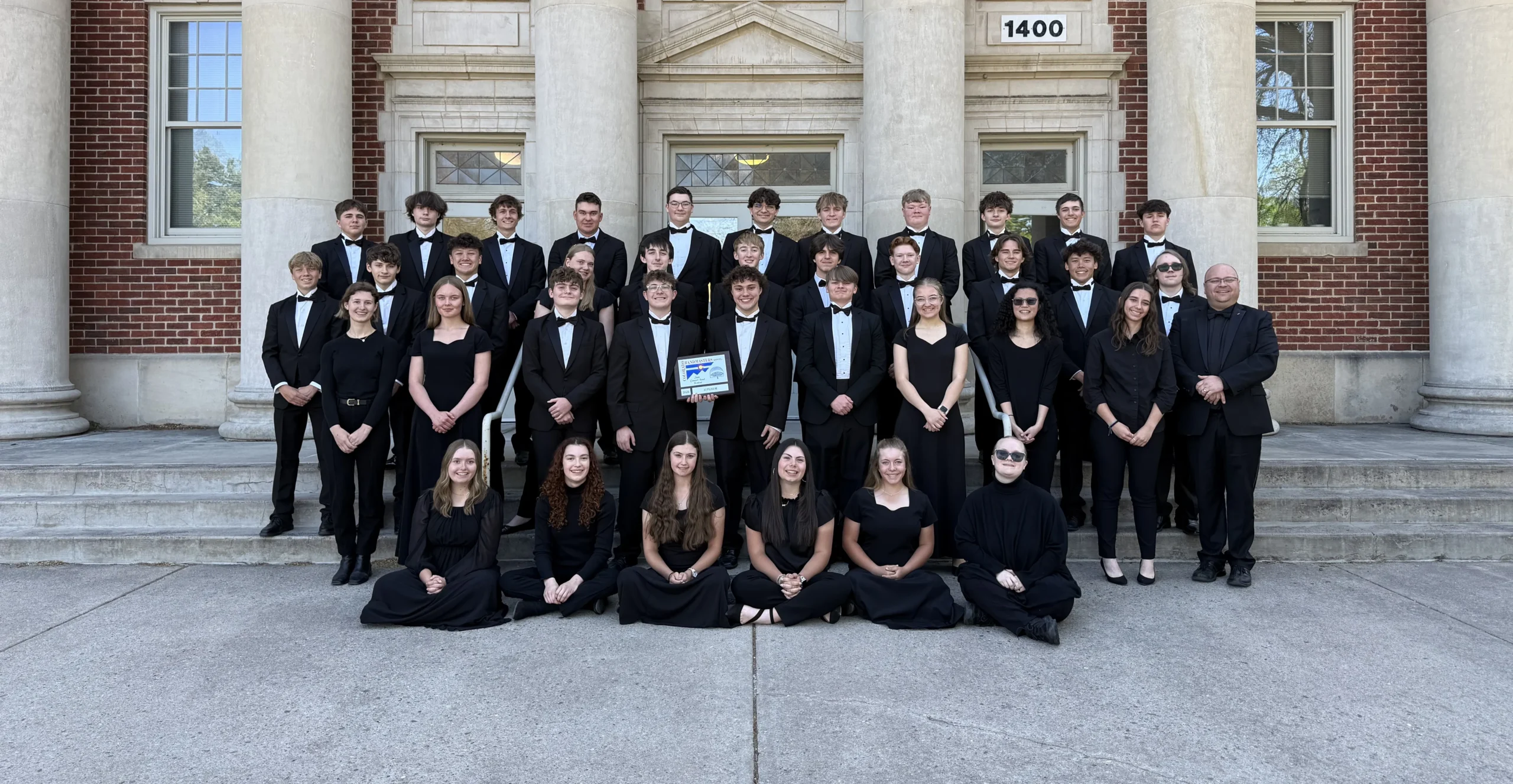 A large choir group in formal black concert attire poses on the steps of a brick building with stone columns.